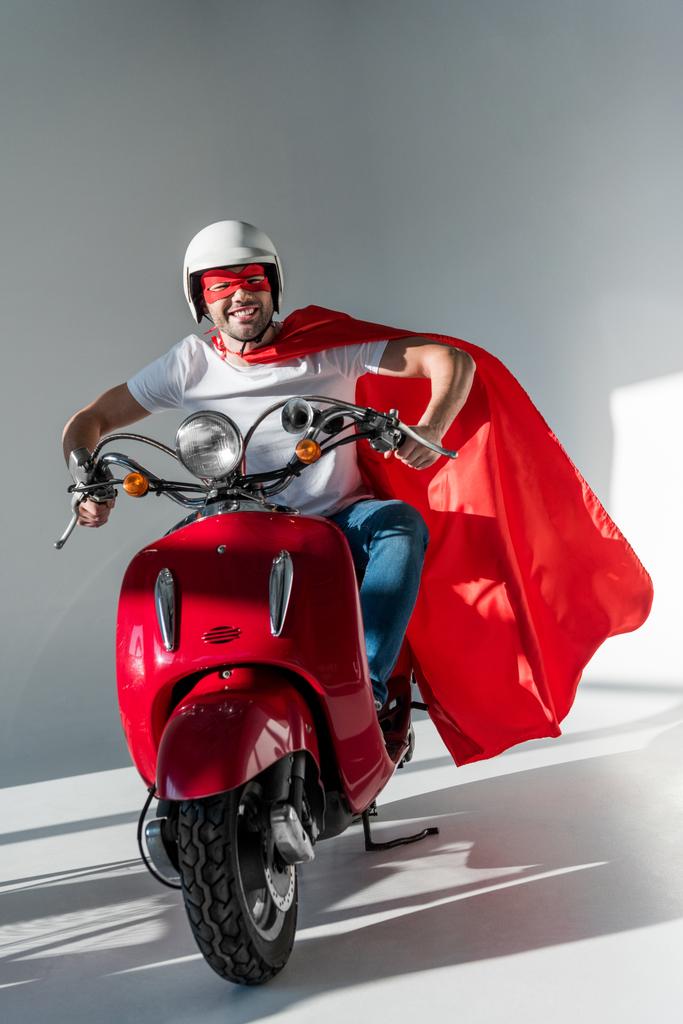 smiling man in protective helmet, superhero mask and cape riding red scooter - Photo, Image