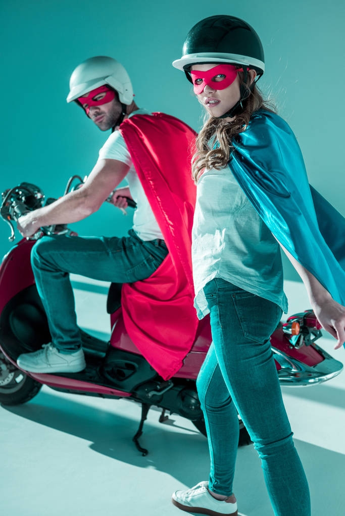 stijlvolle paar in helmen en superheld kostuums met rode scooter camera kijken - Foto, afbeelding