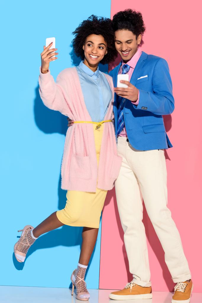 Jonge Afrikaanse amercian meisje dat neemt selfie en knuffelen stijlvolle man die naar zijn telefoon op roze en blauwe achtergrond kijkt  - Foto, afbeelding