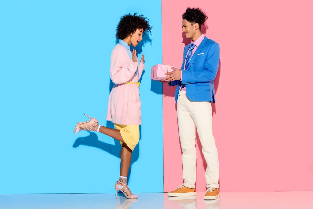 junge afrikanisch-amerikanische Mann gibt Mädchen Geschenk-Box auf rosa und blauen Hintergrund  - Foto, Bild