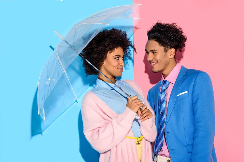 ピンクとブルーの背景の傘の下でお互いに近い笑みを浮かべてカップルの立っています。   - 写真・画像