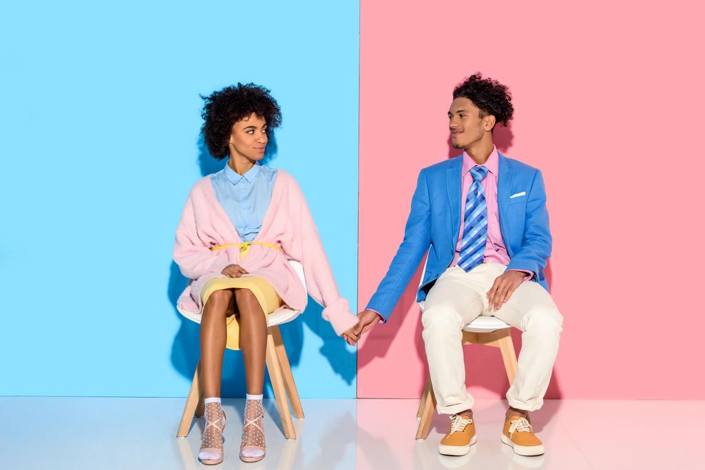 Африканская американская пара, держась за руки, сидя на стульях на розовом и синем фоне
 - Фото, изображение