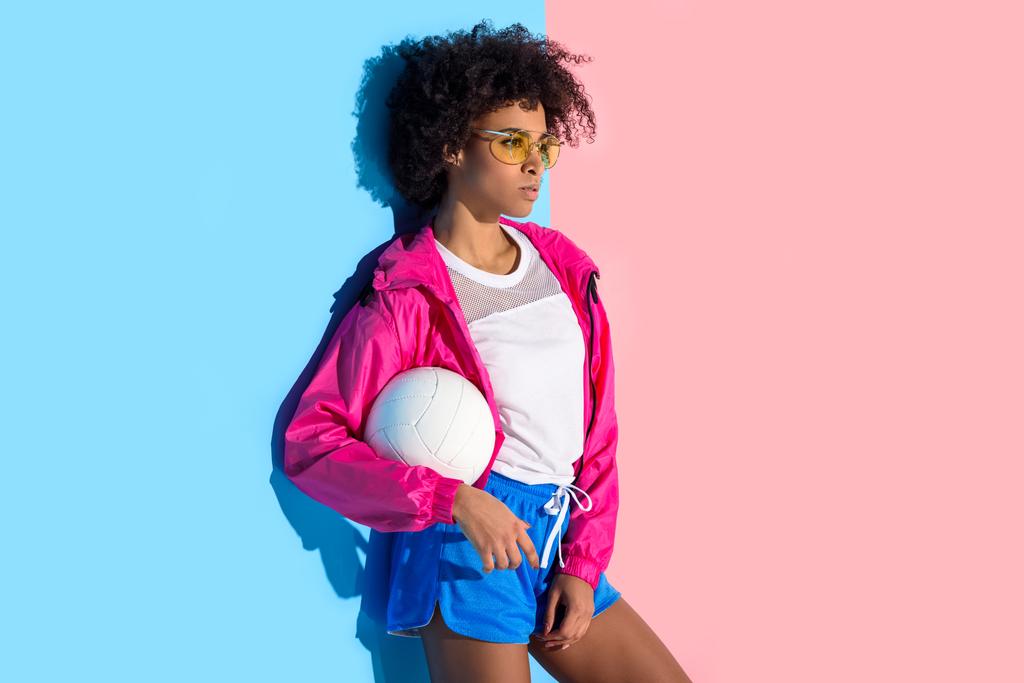  Young girl in glasses holding  ball and looking at away on pink and blue background  - Photo, Image