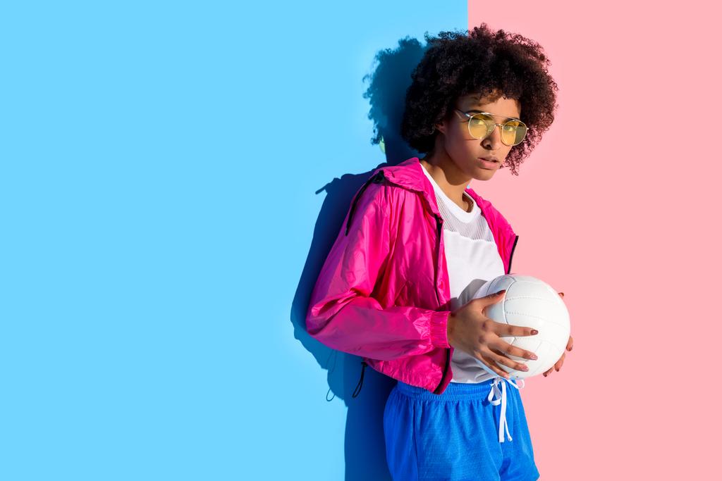  Fiatal lány szemüvegét, tartja a labdát és látszó-on fényképezőgép-rózsaszín és a kék háttér  - Fotó, kép
