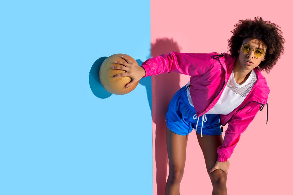 Chica joven en gafas sosteniendo pelota de baloncesto y mirando hacia otro lado sobre fondo rosa y azul
   - Foto, Imagen