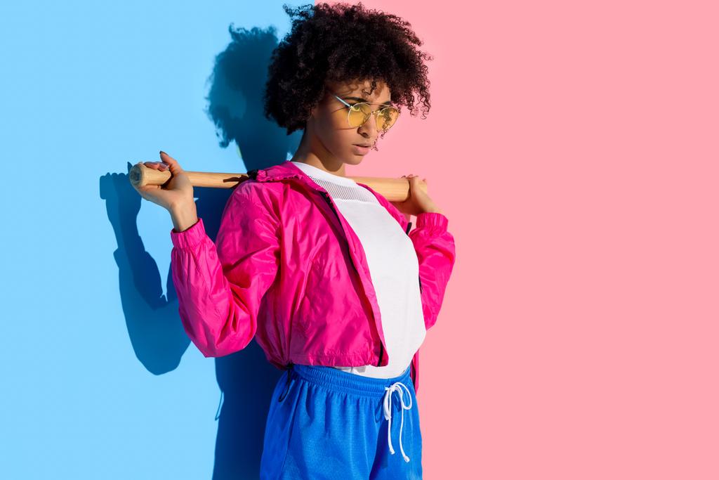 Fiatal világos afro-amerikai lány baseball ütővel a vállán, a rózsaszín és a kék háttér  - Fotó, kép