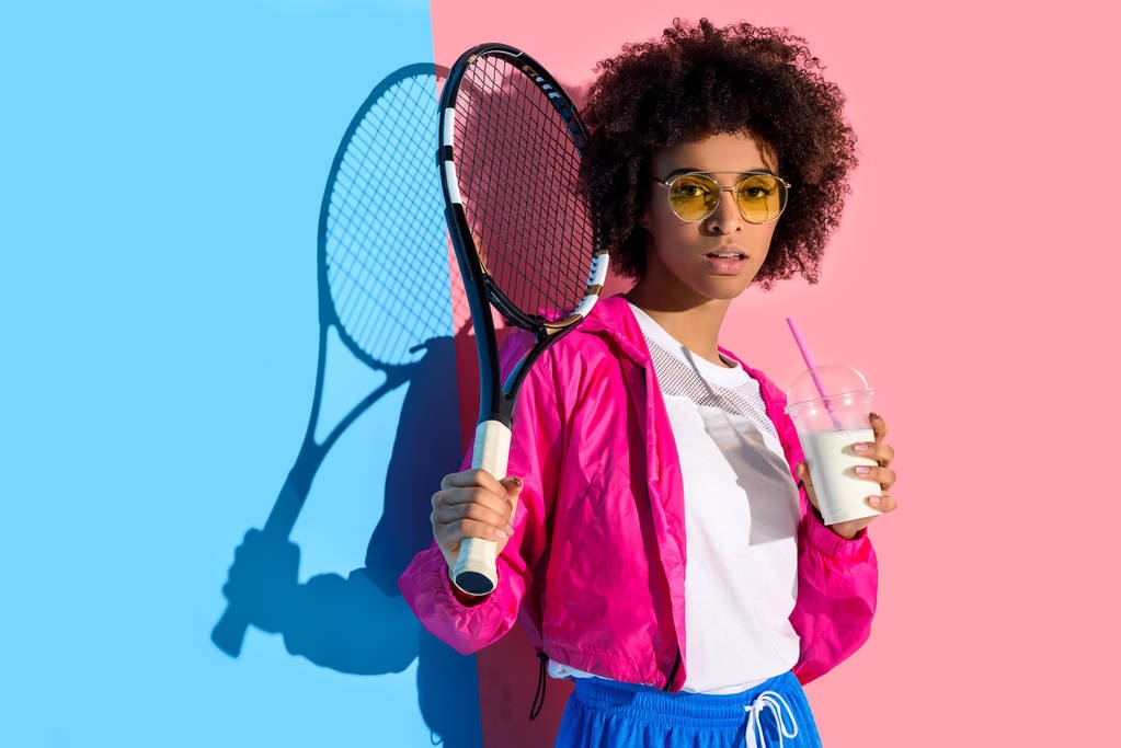 ピンクとブルーの背景にテニス ラケット、ドリンクをプラスチック製のカップを保持している明るいアフリカ系アメリカ人少女  - 写真・画像