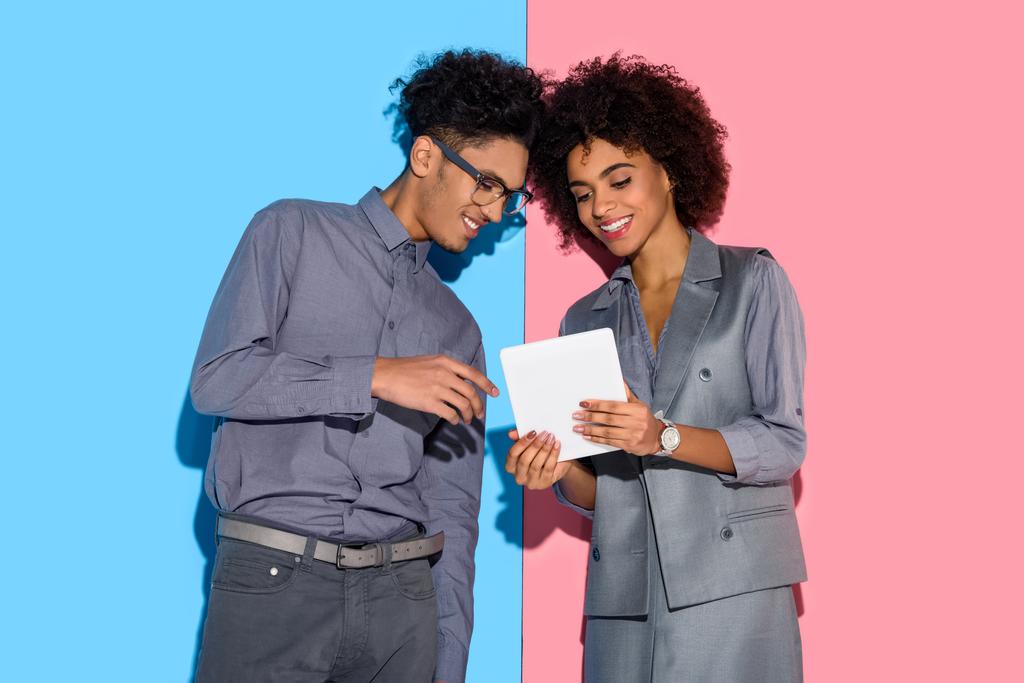 junge afrikanisch-amerikanische Geschäftsfrau und Geschäftsfrau schaut in Tablette in den Händen auf rosa und blauem Hintergrund  - Foto, Bild