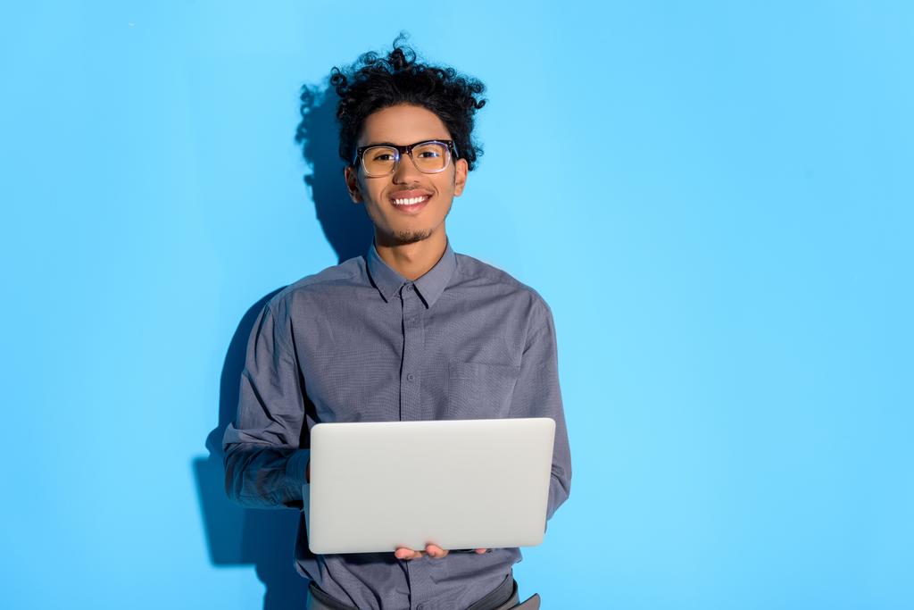 Молодий афроамериканський посміхається чоловік тримає ноутбук на синьому фоні
  - Фото, зображення