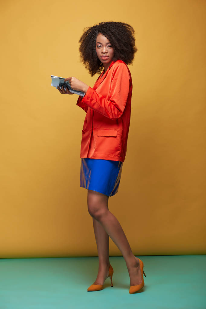 Színes portré fiatal afrikai lány afro frizura. Lány visel narancssárga kabátot copybook tartja a kezét, és pózol a sárga háttéren. Stúdió felvétel. - Fotó, kép
