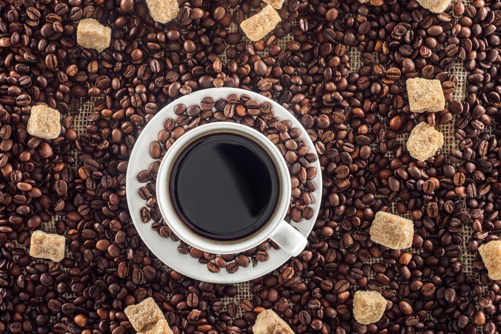 Tasse Kaffee mit Untertasse, gerösteten Kaffeebohnen und braunem Zucker auf Sacktuch - Foto, Bild