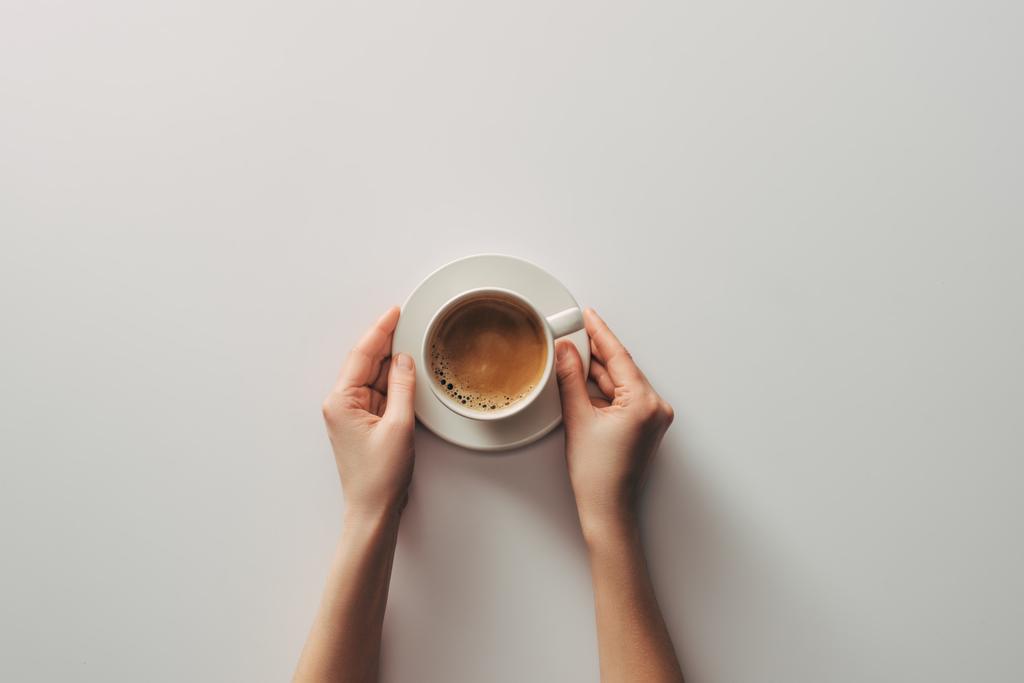 Draufsicht auf weibliche Hände und Tasse Kaffee mit Untertasse auf grau - Foto, Bild