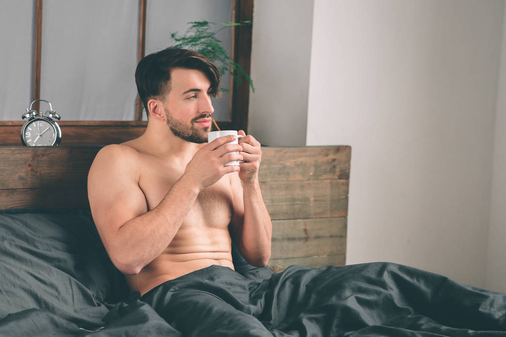 schöner lächelnder junger nackter Mann, der eine Tasse Kaffee in der Hand hält und wegschaut, während er auf dem Bett sitzt - Foto, Bild