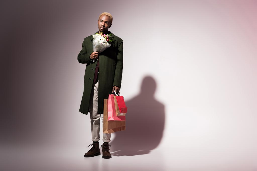 Стильный молодой африканский американец держит букет цветов и яркие сумки для покупок
 - Фото, изображение