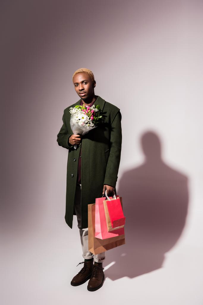 花と明るいショッピング バッグの花束を保持しているスタイリッシュな若いアフリカ系アメリカ人 - 写真・画像