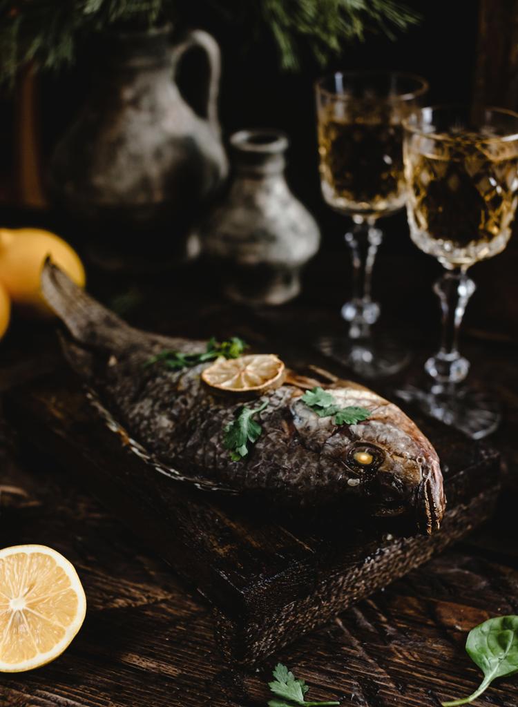 gebackener Fisch mit Zitrone und Kräutern auf Holzbrett mit Weißweingläsern auf dunklem Holztisch - Foto, Bild
