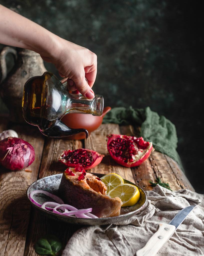 weibliche Hand gießt Öl über gebackenes Lachssteak mit Zitrone und Zwiebel auf Teller mit Granatapfel auf Holztisch - Foto, Bild