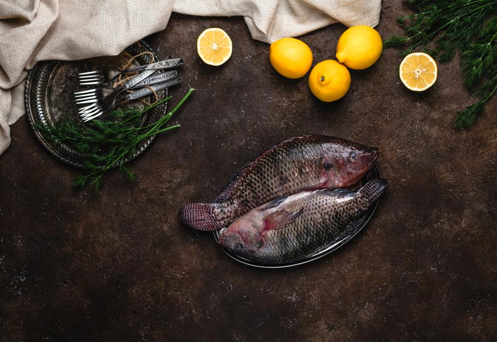 κάτοψη του ακατέργαστου υγιή ψάρια στο πιάτο και τα λεμόνια με βότανα σε σκούρο επιτραπέζια - Φωτογραφία, εικόνα