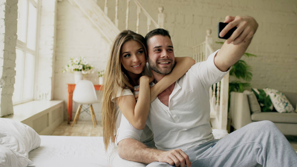 Fiatal, szép és szerető pár fényképet selfie a smartphone kamera miközben ül az ágyban reggel - Fotó, kép
