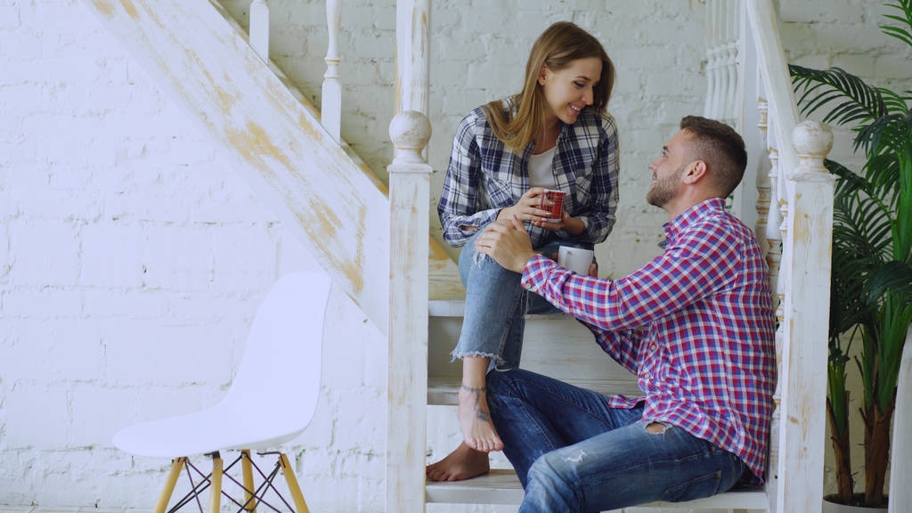 Młoda para szczęśliwy, a kochający pić herbatę i rozmowy siedząc na schodach w domu salon. - Zdjęcie, obraz