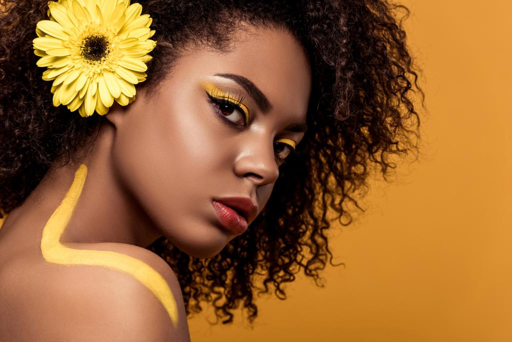 Joven mujer afroamericana tierna con maquillaje artístico y gerbera en el pelo mirando a la cámara aislada sobre fondo naranja
 - Foto, imagen