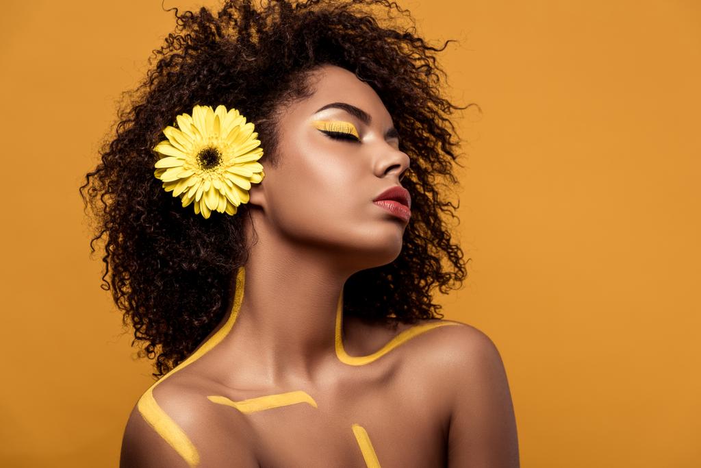 Aantrekkelijke jonge african american vrouw met artistieke make-up en gerbera in haar geïsoleerd op een oranje achtergrond - Foto, afbeelding