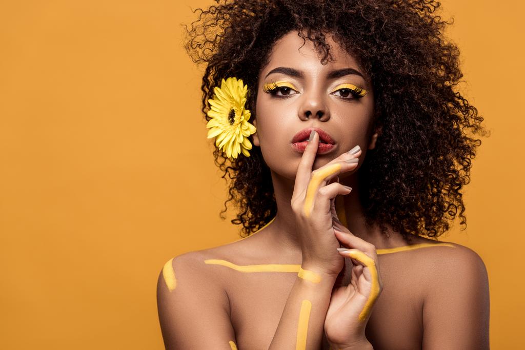 Jonge sensuele african american vrouw met artistieke make-up en gerbera in haar vinger te houden op lippen geïsoleerd op een oranje achtergrond - Foto, afbeelding