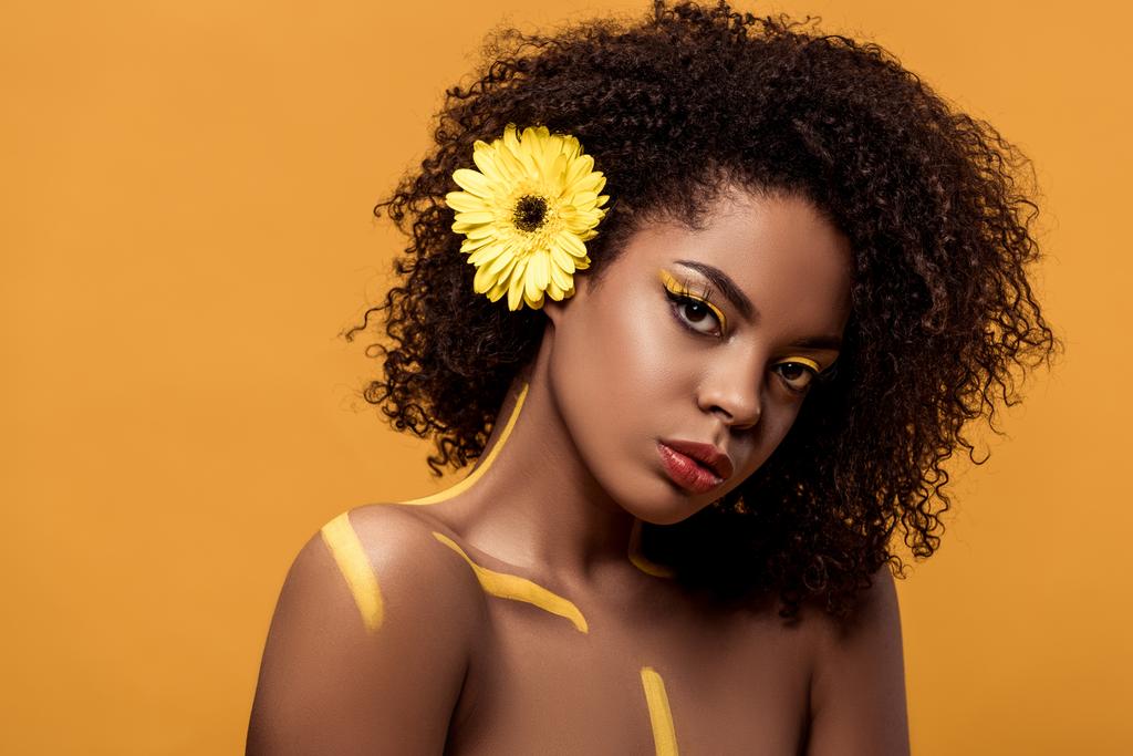 Молодая ярко-африканская американка с художественным макияжем и герберой в волосах, изолированных на оранжевом фоне
 - Фото, изображение