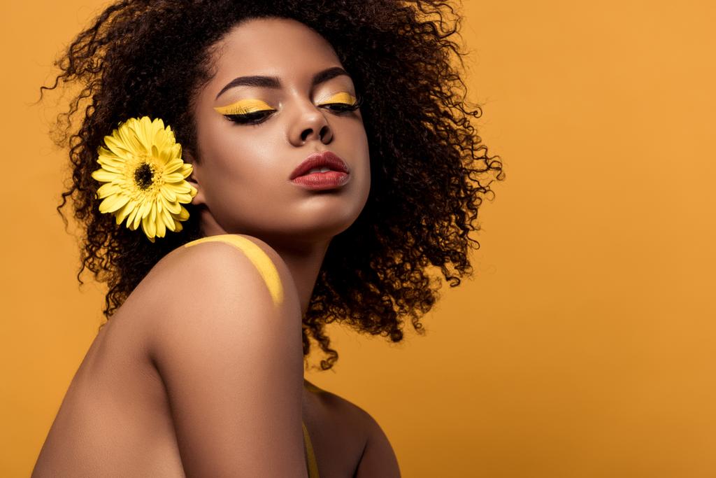 Mujer afroamericana tierna joven con maquillaje artístico y gerbera en pelo aislado sobre fondo naranja
 - Foto, Imagen