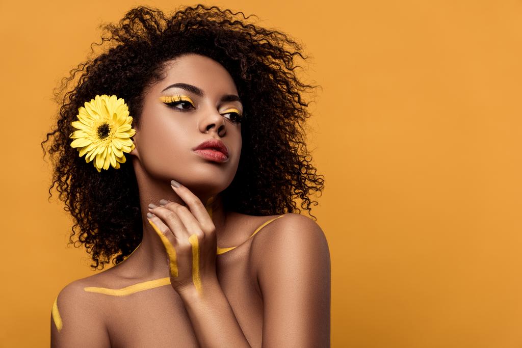 Mladá žena smyslné afroamerické umělecký make-up a gerbera ve vlasech drží ruku z její tváře izolované na oranžové pozadí - Fotografie, Obrázek