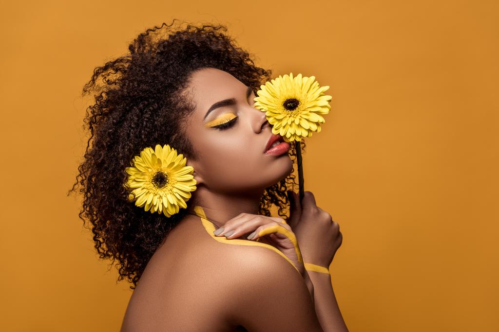 Elegante mujer afroamericana con maquillaje artístico sostiene la flor por su cara toca tiernamente su piel aislada sobre fondo naranja
 - Foto, imagen