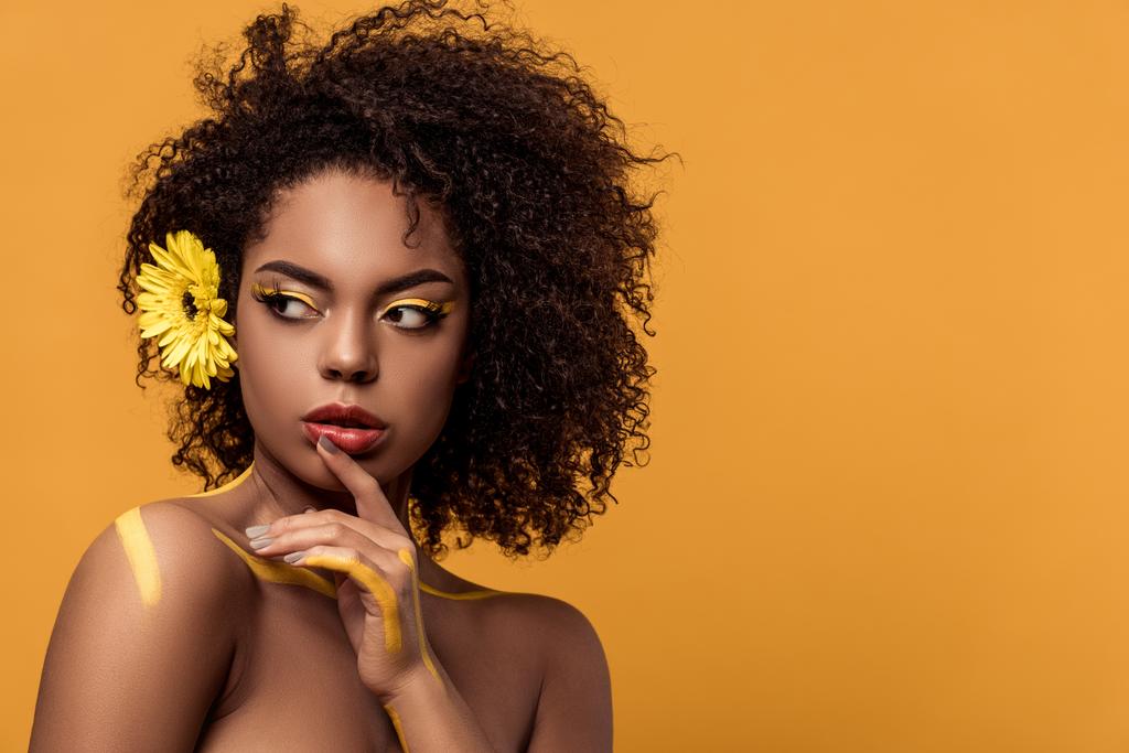 Joven mujer afroamericana brillante con maquillaje artístico y gerbera en el pelo sostiene la mano por su cara aislada sobre fondo naranja
 - Foto, Imagen