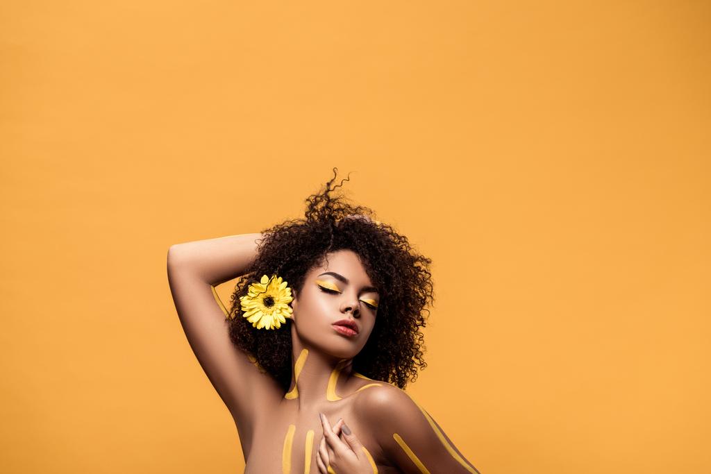 Young sensual african american woman with artistic make-up and gerbera in hair, arm behind her head isolated on orange background - Photo, Image