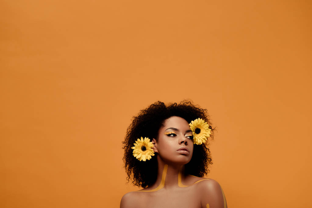 Joven mujer afroamericana sensual con maquillaje artístico y gerbera en pelo aislado sobre fondo naranja
 - Foto, Imagen