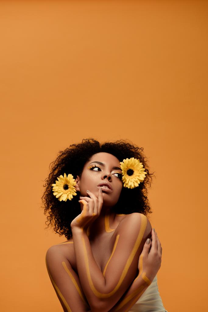 Jeune femme africaine américaine sensuelle avec maquillage artistique et gerbera dans les cheveux isolés sur fond orange
 - Photo, image