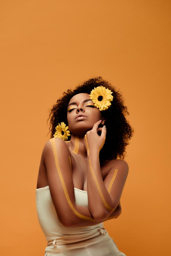 Genç Afro-Amerikan kadın sanatsal makyaj ve saç üzerinde turuncu arka plan izole gerbera ile ihale - Fotoğraf, Görsel