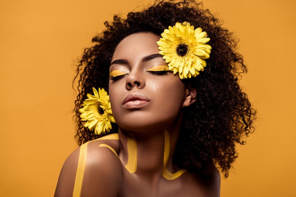 Young sensual african american woman with artistic make-up and gerbera in hair isolated on orange background - Photo, Image