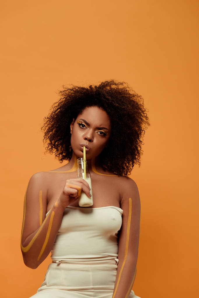 Jovem americana africana pensativa com maquiagem artística bebendo leite de garrafa isolada no fundo laranja
 - Foto, Imagem