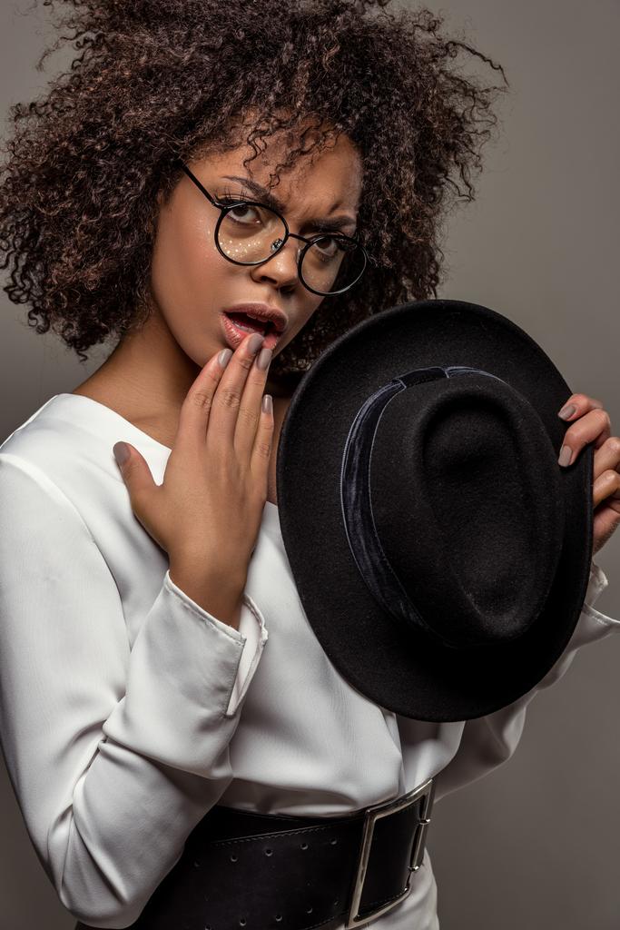 Young perplexed african american woman in white shirt wearing glasses and holding hat isolated on grey background - Photo, Image