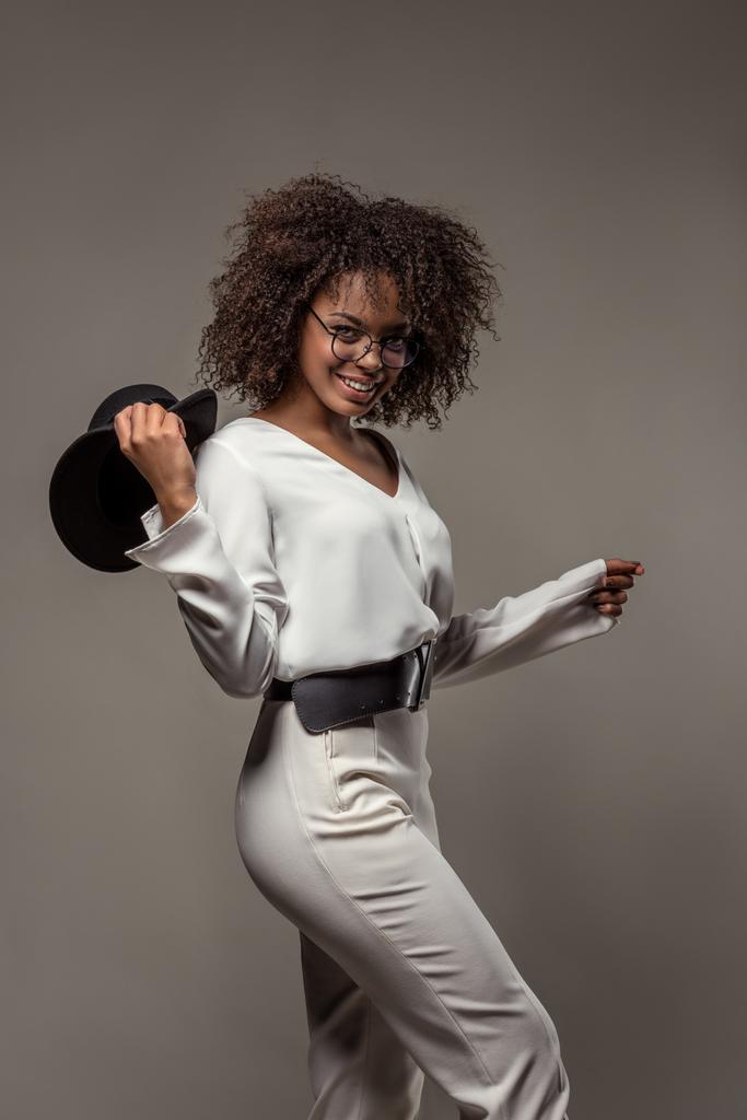 Stylish african american woman in white clothes smiling and waving black hat isolated on grey background - Photo, Image