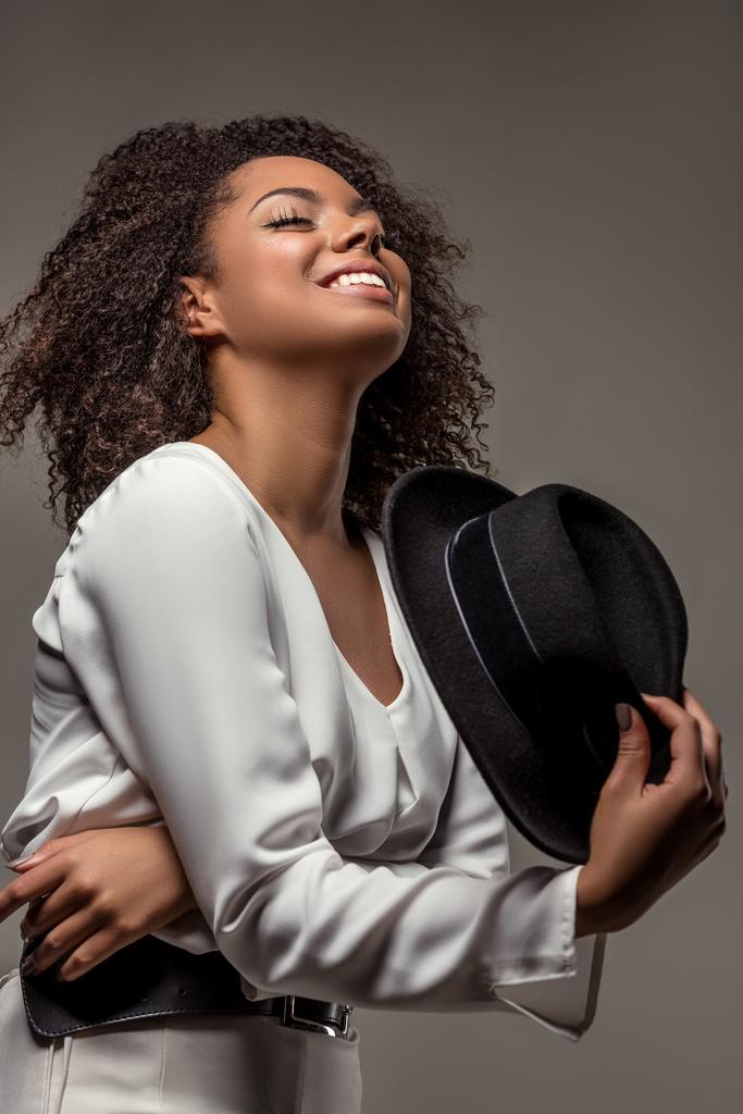 Femme afro-américaine élégante en vêtements blancs souriant et agitant chapeau noir isolé sur fond gris
 - Photo, image
