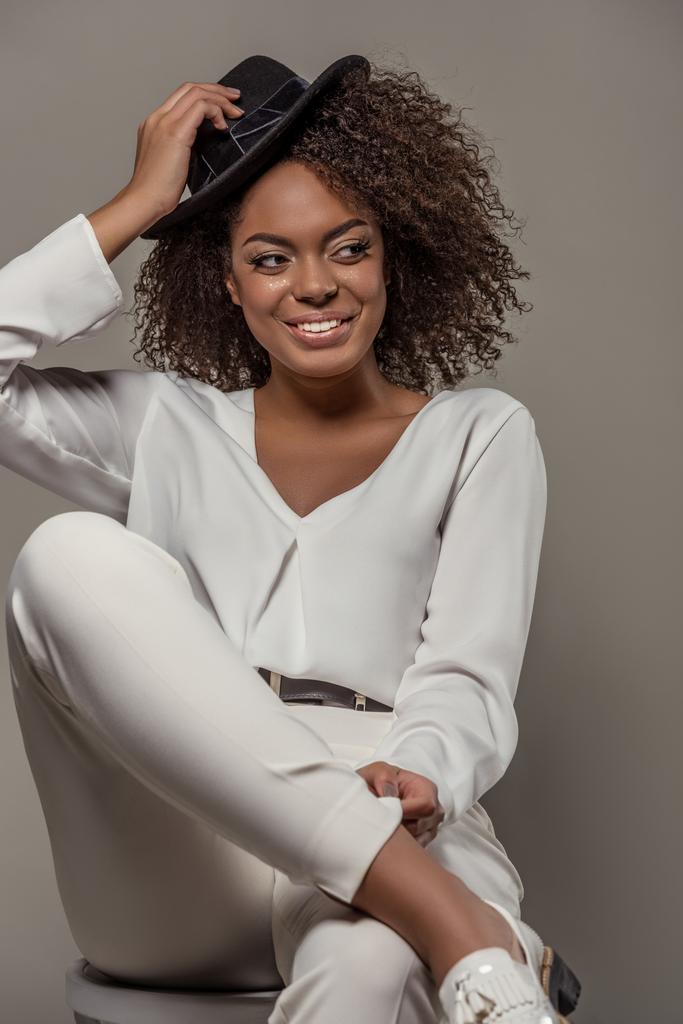 Beautiful african american woman in white clothes smiling and holding black hat isolated on grey background - Photo, Image
