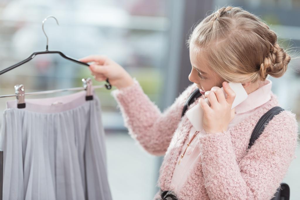 dziecko mówiąc na smartphone, trzymając w ręku tkaniny na wieszaku w sklepie  - Zdjęcie, obraz