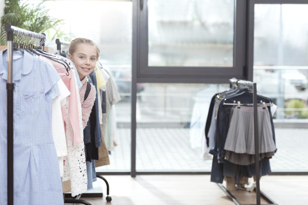 hymyilevä lapsi etsii ulos rivi vaatteita ripustin kaupassa
   - Valokuva, kuva