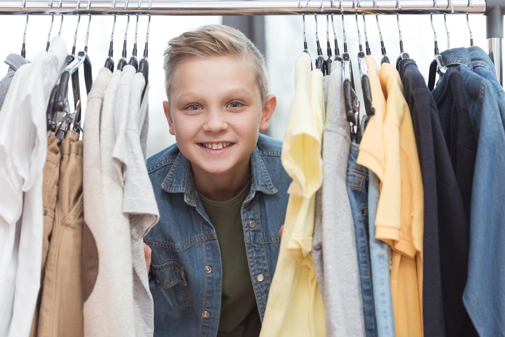 Lächelnder Junge blickt in die Kamera, umgeben von Kleidern auf Kleiderbügel im Geschäft   - Foto, Bild