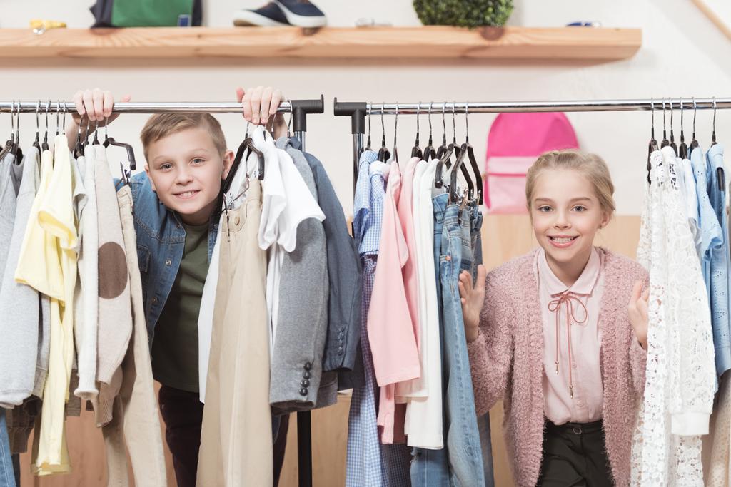 two smiling kids looking at camera while standing under cloth hanger at shop  - Photo, Image