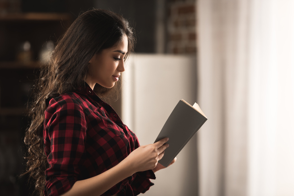 Zijaanzicht van sexy meisje in geruite overhemd leesboek thuis - Foto, afbeelding