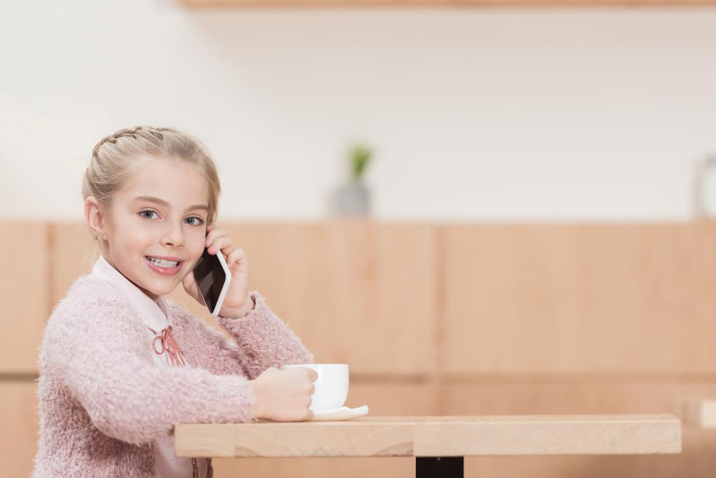 hymyilevä lapsi istuu kuppi käsissä ja käyttää älypuhelinta katsoessaan kameraa
 - Valokuva, kuva