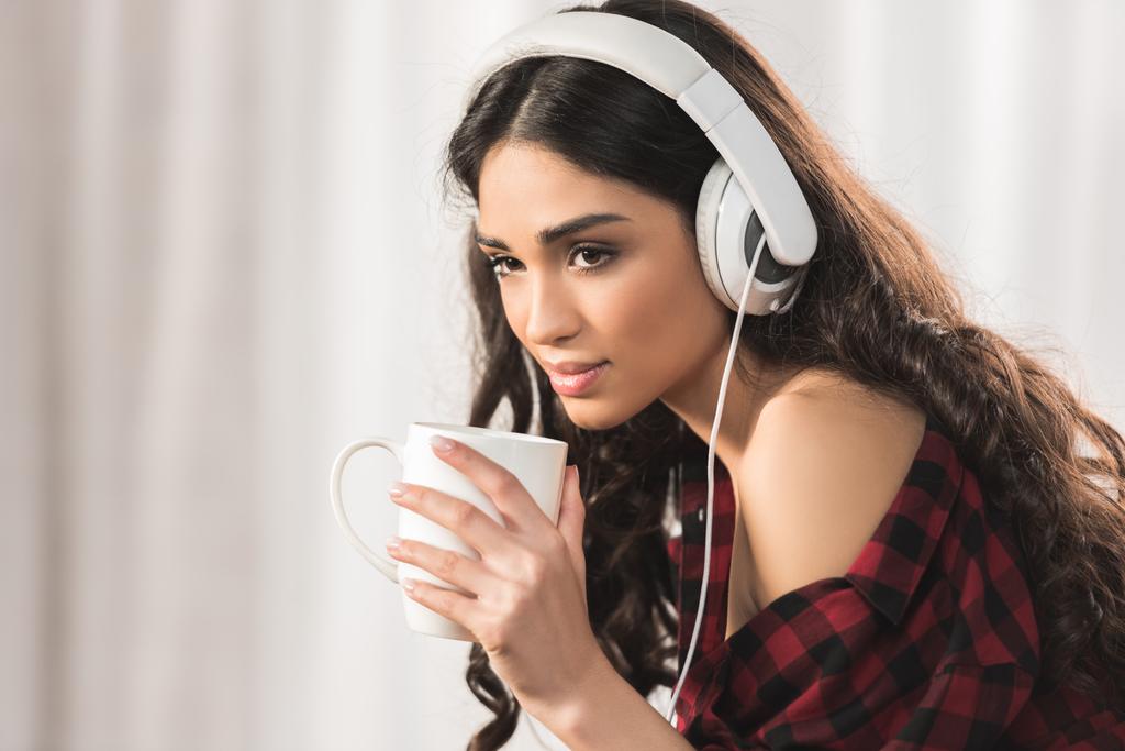 vonzó fiatal nő hallgat zenét a fejhallgatót, és kávét iszik, otthon - Fotó, kép