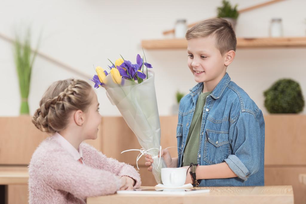 χαμογελαστό αγόρι παρουσιάζοντας λουλούδια σε κορίτσι που κάθεται στο τραπέζι φίλη  - Φωτογραφία, εικόνα